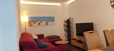 Wohnzimmer mit rotem Sofa, Fernseher, Couchtisch und Landschaftsbild an der Wand.